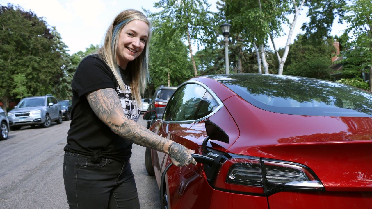 On a residential city street, a woman with a tattooed arm stands next to a red Tesla, holding the car's charging cable in her right hand while she smiles at the camera. 