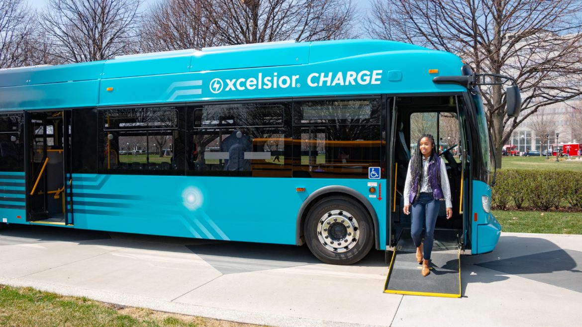 An xcelsior Charge bus, with green and black livery, is parked on a city street. A young woman walks down the bus' ramp.