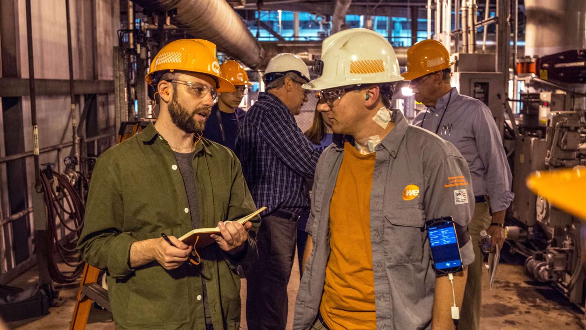 In a factory-like environment, several men wearing hard hats stand around, discussing things.
