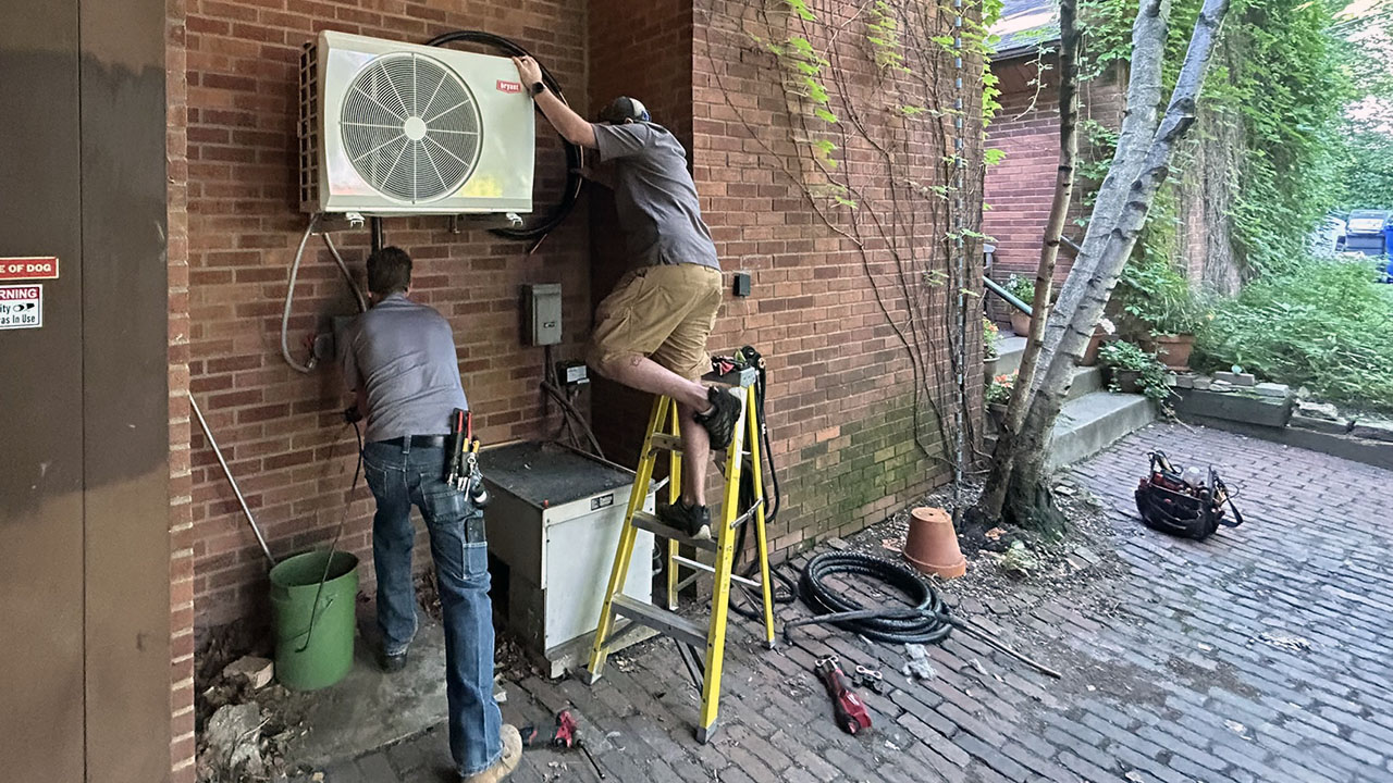 Two workers installing heat pump on the side of a brick home.