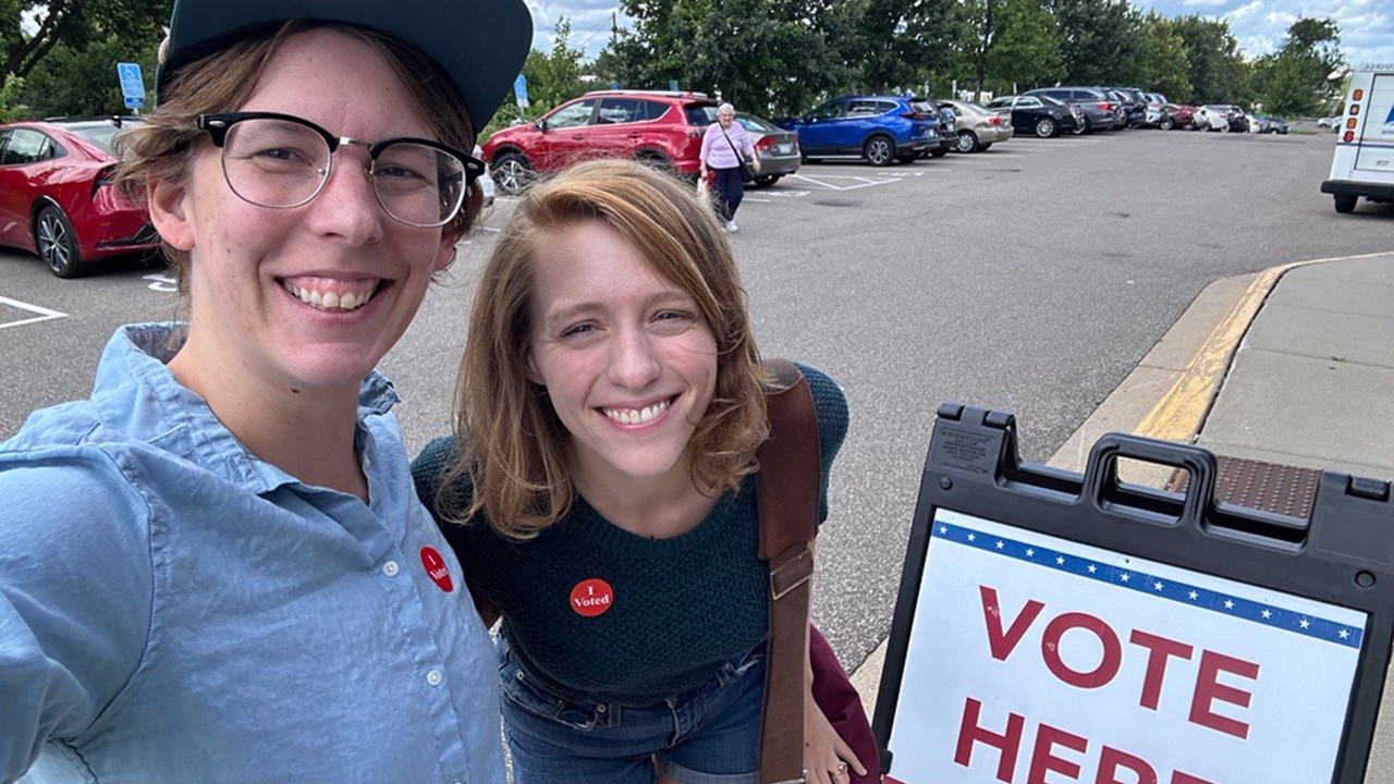 Two people smiling in front of sign that says "Vote Here" 