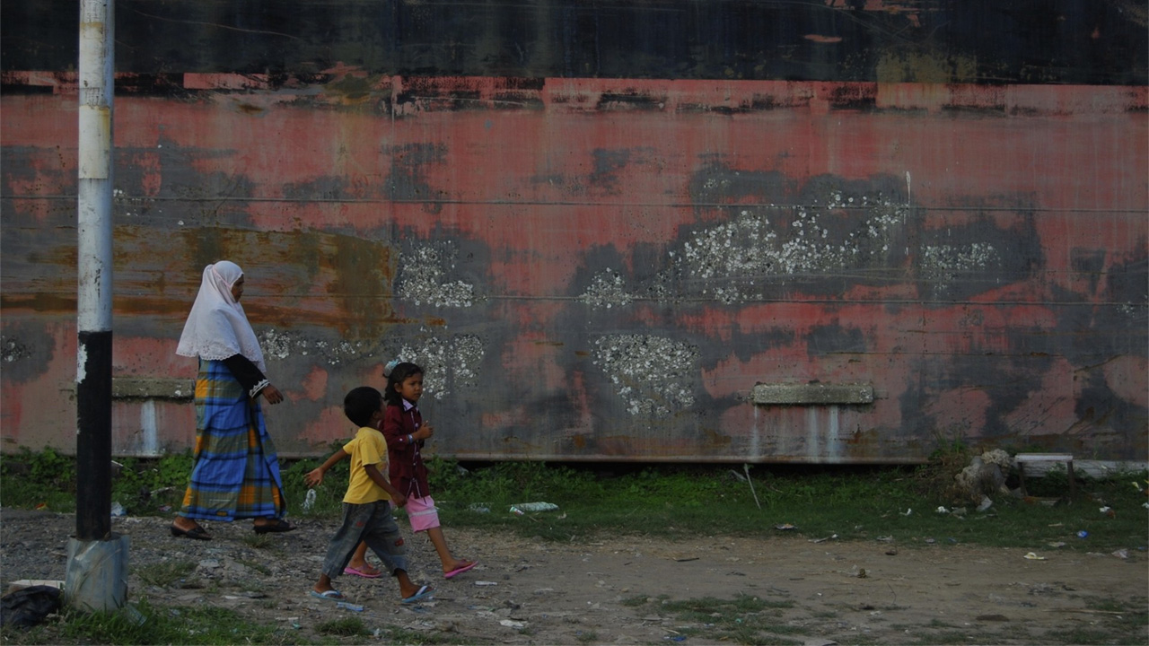 A mother walks behind her two kids past a red oil barge.
