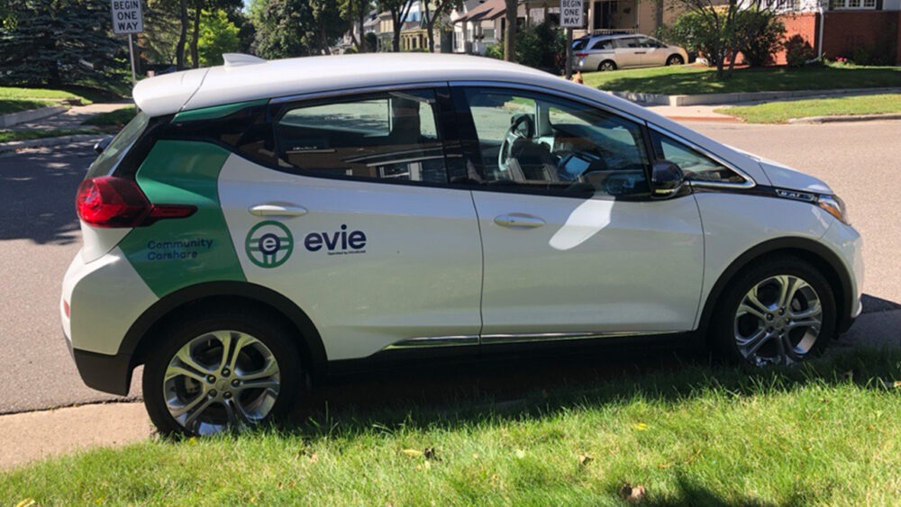 A white Evie vehicle parked on a Minnesota street in summer.