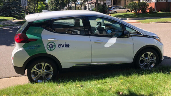 A white Evie vehicle parked on a Minnesota street in summer.
